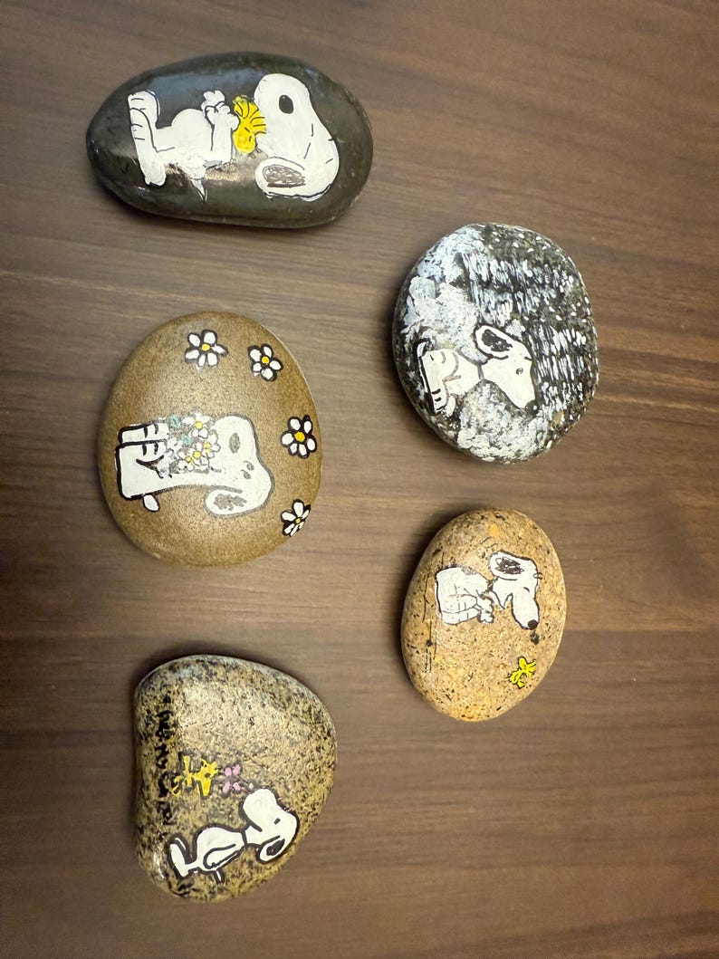 Puede incluir: Cinco piedras pintadas con Snoopy de la tira c&oacute;mica Peanuts. Las piedras son de forma ovalada y est&aacute;n pintadas con detalles blancos y amarillos sobre un fondo oscuro. Algunas piedras tienen a Snoopy con flores.