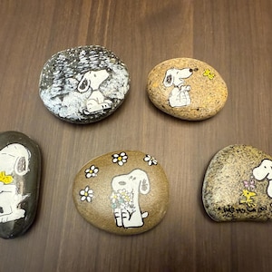 Puede incluir: Cinco piedras pintadas a mano con Snoopy de Peanuts. Cada piedra tiene un dise&ntilde;o diferente, incluyendo a Snoopy con flores, Snoopy con un p&aacute;jaro y Snoopy en una escena invernal. Las piedras est&aacute;n sobre una superficie de madera marr&oacute;n.