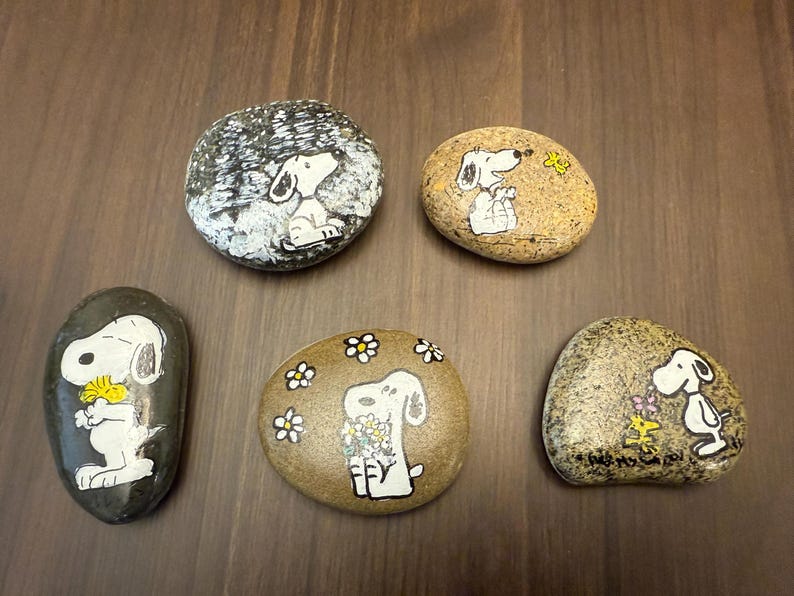 Puede incluir: Cinco piedras pintadas a mano con Snoopy de Peanuts. Cada piedra tiene un dise&ntilde;o diferente, incluyendo a Snoopy con flores, Snoopy con un p&aacute;jaro y Snoopy en una escena invernal. Las piedras est&aacute;n sobre una superficie de madera marr&oacute;n.