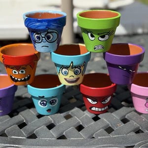 May include: A collection of small, hand-painted terracotta flower pots. Each pot is painted a different color and features a cartoon face with various expressions, including sadness, anger, and joy. The pots are stacked on a dark gray surface.