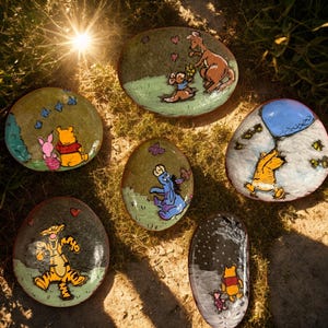 May include: A collection of six painted stones featuring characters from Winnie the Pooh. The stones are painted with scenes of Pooh, Piglet, Tigger, Eeyore, Kanga, and Roo. The stones are on a bed of grass.