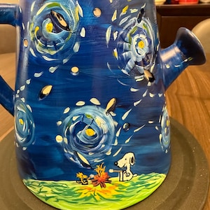 May include: A hand-painted blue ceramic watering can featuring a starry night design with white and yellow brushstrokes. The base depicts a cartoon dog sitting by a campfire. The watering can has a handle and a spout.