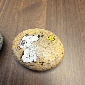 Puede incluir: Piedra ovalada pintada a mano con Snoopy de la tira c&oacute;mica Peanuts. Snoopy est&aacute; representado en blanco y negro, mirando a un p&aacute;jaro amarillo. La piedra tiene una superficie moteada y de color beige.