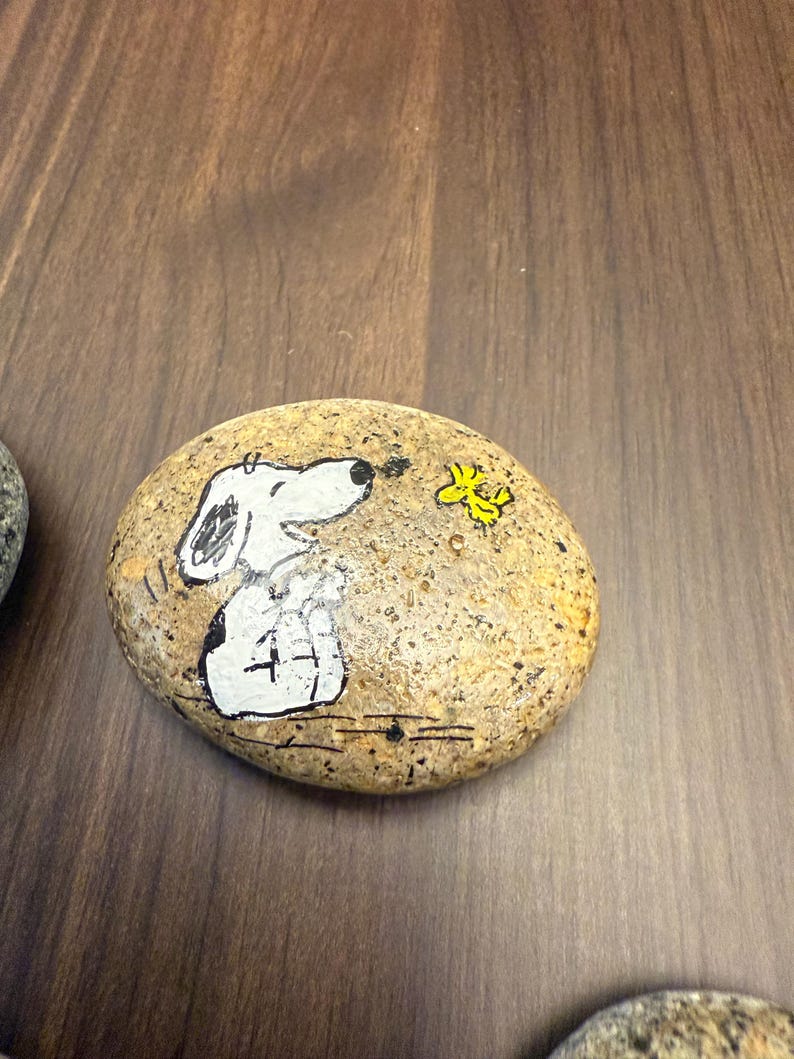 Puede incluir: Piedra ovalada pintada a mano con Snoopy de la tira c&oacute;mica Peanuts. Snoopy est&aacute; representado en blanco y negro, mirando a un p&aacute;jaro amarillo. La piedra tiene una superficie moteada y de color beige.