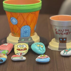 May include: Hand-painted flower pots and decorative rocks featuring characters from the animated series. The orange pot is designed to resemble a pineapple house, while the other pot is gray with the text "CHUM BUCKET". The rocks are painted with various character faces.
