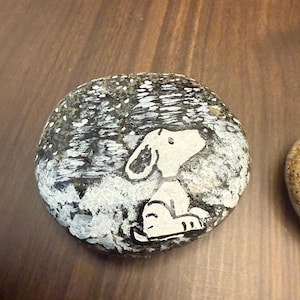 Puede incluir: Piedra pintada a mano con un perro de dibujos animados blanco sobre un fondo nevado. El perro est&aacute; sentado y mirando hacia arriba. La piedra est&aacute; sobre una superficie de madera. El fondo tiene un efecto moteado.