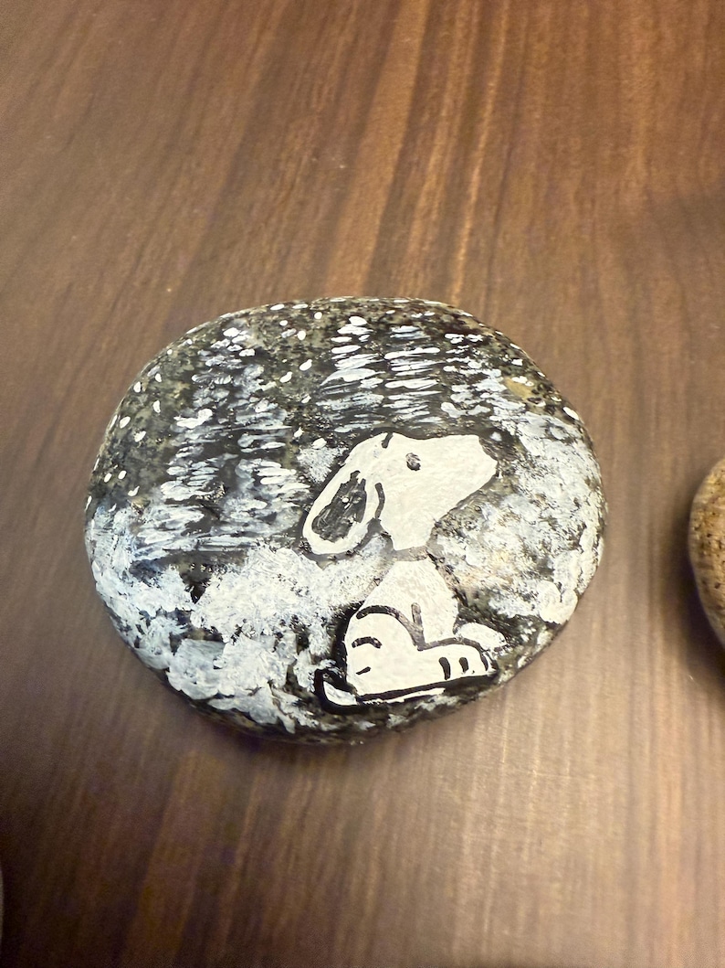 Puede incluir: Piedra pintada a mano con un perro de dibujos animados blanco sobre un fondo nevado. El perro est&aacute; sentado y mirando hacia arriba. La piedra est&aacute; sobre una superficie de madera. El fondo tiene un efecto moteado.