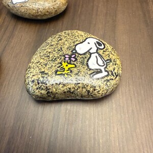 Puede incluir: Dos piedras pintadas con los personajes de Snoopy y Woodstock. Una piedra muestra a Snoopy sosteniendo una flor, y la otra muestra a Snoopy con una flor. Las piedras est&aacute;n pintadas con detalles en negro, blanco, amarillo y rosa.