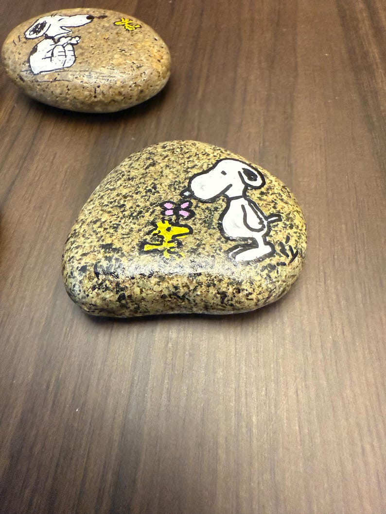 Puede incluir: Dos piedras pintadas con los personajes de Snoopy y Woodstock. Una piedra muestra a Snoopy sosteniendo una flor, y la otra muestra a Snoopy con una flor. Las piedras est&aacute;n pintadas con detalles en negro, blanco, amarillo y rosa.