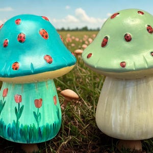 May include: Two decorative mushroom figurines. One is teal with red ladybug spots and painted tulips. The other is light green with red spots. Both have cream-colored stems and are set in a grassy field.