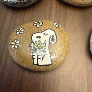 Puede incluir: Piedra pintada a mano con un perro de dibujos animados blanco sosteniendo un ramo de flores. El perro est&aacute; rodeado de margaritas blancas y amarillas. La piedra es de color marr&oacute;n claro y est&aacute; sobre una superficie de madera.
