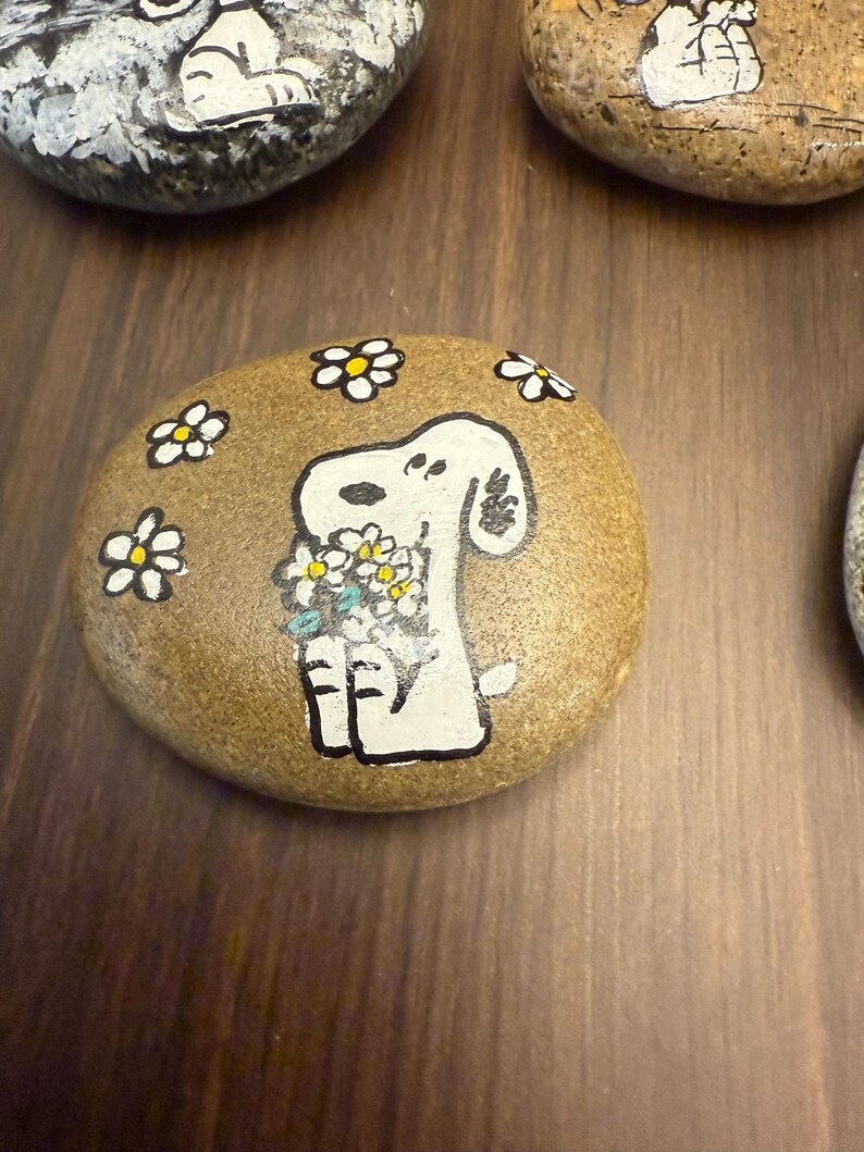 Puede incluir: Piedra pintada a mano con un perro de dibujos animados blanco sosteniendo un ramo de flores. El perro est&aacute; rodeado de margaritas blancas y amarillas. La piedra es de color marr&oacute;n claro y est&aacute; sobre una superficie de madera.