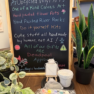 May include: A chalkboard sign with the text "Art by Josie" lists handmade items like upcycled vinyl art, canvas art, flower pots, and DIY kits. A small canvas, paint set, and flower pot are displayed in front of the sign.