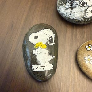 Puede incluir: Piedras pintadas a mano con Snoopy de Peanuts. Una piedra muestra a Snoopy sosteniendo una flor amarilla, otra muestra a Snoopy sentado y una tercera tiene flores blancas. Las piedras est&aacute;n sobre una superficie marr&oacute;n.