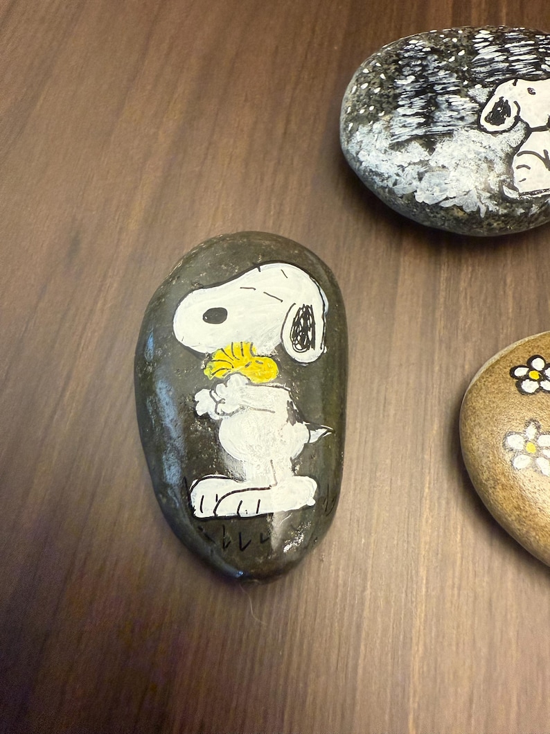 Puede incluir: Piedras pintadas a mano con Snoopy de Peanuts. Una piedra muestra a Snoopy sosteniendo una flor amarilla, otra muestra a Snoopy sentado y una tercera tiene flores blancas. Las piedras est&aacute;n sobre una superficie marr&oacute;n.