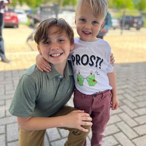 May include: Two young boys are smiling and hugging. The boy in the green shirt is kneeling and has his arm around the boy in the white shirt. The boy in the white shirt is wearing a shirt that says "PROST!" with a picture of two juice boxes on it.