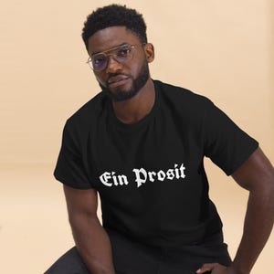 May include: Black t-shirt with the white text "Ein Prosit" in a gothic font. The shirt is made of a soft material and is suitable for casual wear. The t-shirt is a classic crew neck style.