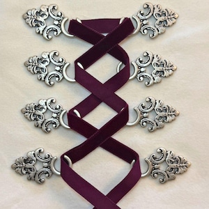May include: Close-up of a decorative garment closure. Four ornate silver-toned clasps are connected by a deep purple velvet ribbon, creating a crisscross pattern. The clasps have a detailed, vintage-inspired design.