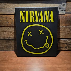May include: Black canvas art featuring the word "NIRVANA" in yellow, above a yellow smiley face with crossed-out eyes and a protruding tongue. The artwork is displayed against a wooden background.
