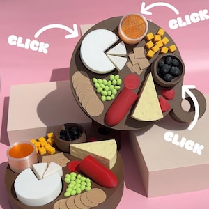 Pode incluir: Uma tábua de servir circular marrom com vários alimentos, incluindo queijo, biscoitos, azeitonas e uma salsicha vermelha. A tábua está disposta sobre um fundo rosa com a palavra "CLICK" em branco.