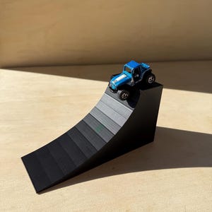 May include: A small, blue toy jeep with a white stripe sits atop a black ramp. The ramp has a gradient design, transitioning from dark to light gray. The toy car has black wheels and a white stripe.