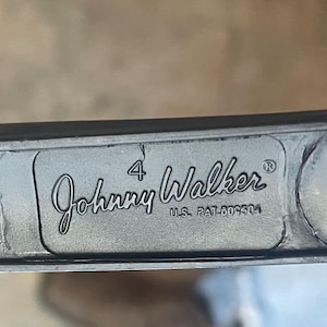 May include: Close-up of a dark gray metal object with the text "Johnny Walker" in a cursive font. The number "4" is above the name, and "U.S. PAT.009304" is below. The object has a circular hole and a circular indentation.