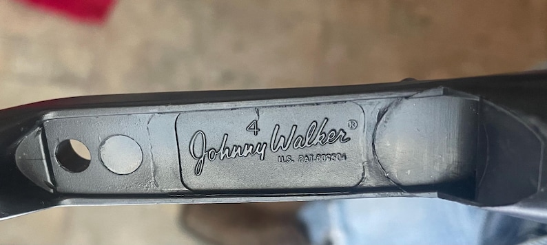 May include: Close-up of a dark gray metal object with the text "Johnny Walker" in a cursive font. The number "4" is above the name, and "U.S. PAT.009304" is below. The object has a circular hole and a circular indentation.