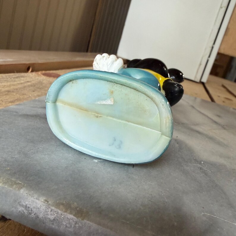 May include: A ceramic figurine base with a light blue oval shape and a visible number 33 stamped on the bottom. The figurine features a black, yellow, and white color scheme with a smooth, glossy finish and a small white paper label attached.