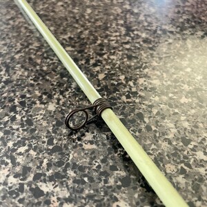 May include: Close-up of a light green fishing rod with a dark metal guide ring. The rod is angled diagonally across the frame, showcasing its smooth texture and the detailed craftsmanship of the guide ring.