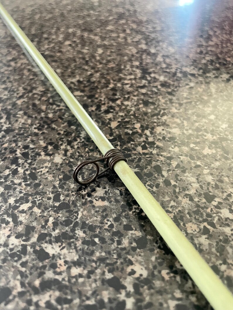 May include: Close-up of a light green fishing rod with a dark metal guide ring. The rod is angled diagonally across the frame, showcasing its smooth texture and the detailed craftsmanship of the guide ring.