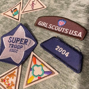 May include: Assortment of Girl Scout patches, including a blue star-shaped patch with "SUPER TROOP GSCC" text, a brown patch with "GIRL SCOUTS USA", and a blue patch with "2004". Various triangular patches are also visible.