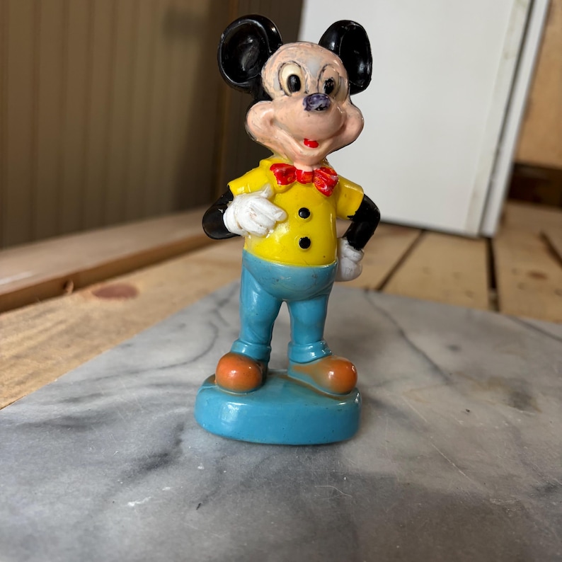May include: A vintage-style Mickey Mouse figurine featuring a pale face, large black ears, and a yellow shirt with a red bowtie. The character wears light blue pants, white gloves, and orange shoes, standing on a solid blue circular base.