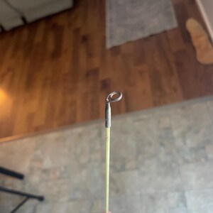 May include: A fishing rod tip with a metal loop at the end. The rod is a light yellow color with a dark gray metal tip. The background is a blurred view of a wooden floor.