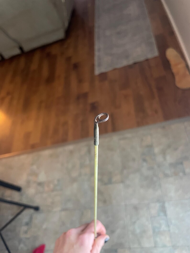 May include: A fishing rod tip with a metal loop at the end. The rod is a light yellow color with a dark gray metal tip. The background is a blurred view of a wooden floor.