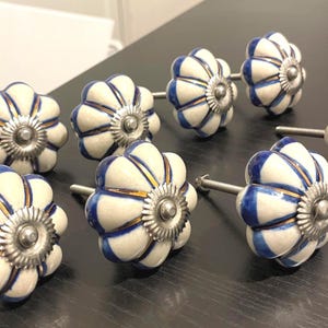 May include: A collection of eight ceramic cabinet knobs. Each knob features a white base with blue and gold accents, and a silver-toned metal base and stem. The knobs are arranged on a dark surface.