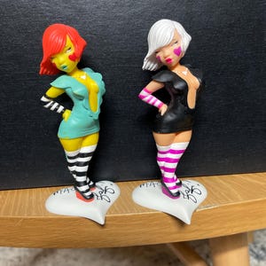 May include: Two figurines of stylized women with red and white hair, wearing dresses and striped socks. One figurine is in a teal dress, the other in a black dress. Both stand on heart-shaped bases.