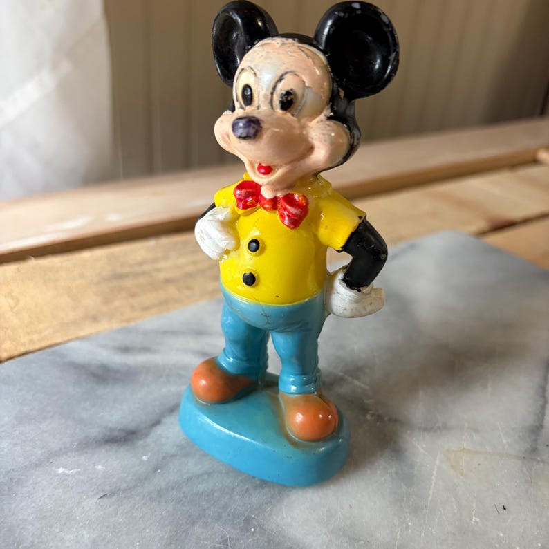 May include: A vintage Mickey Mouse figurine standing on a blue oval base. The character wears a yellow shirt with two black buttons, a red bow tie, blue pants, white gloves, and orange shoes. The figure has black ears and a painted face.