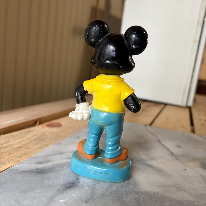 May include: A small figurine of a cartoon mouse character standing on a blue base. The character has a black head with large round ears, a bright yellow short-sleeved shirt, light blue pants, white gloves, and orange shoes. The base has text.