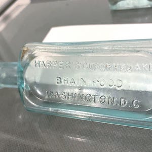 May include: An antique, rectangular, light blue glass bottle. The bottle has a narrow neck and is labeled "HARPER'S SCUROFORHEDAKE BRAIN FOOD WASHINGTON D.C." The bottle is lying on a reflective surface.