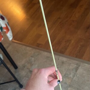 May include: A light green fishing rod with silver hardware. The rod is held at an angle, showing the guides and tip. The background is a wooden floor and a person's arm.