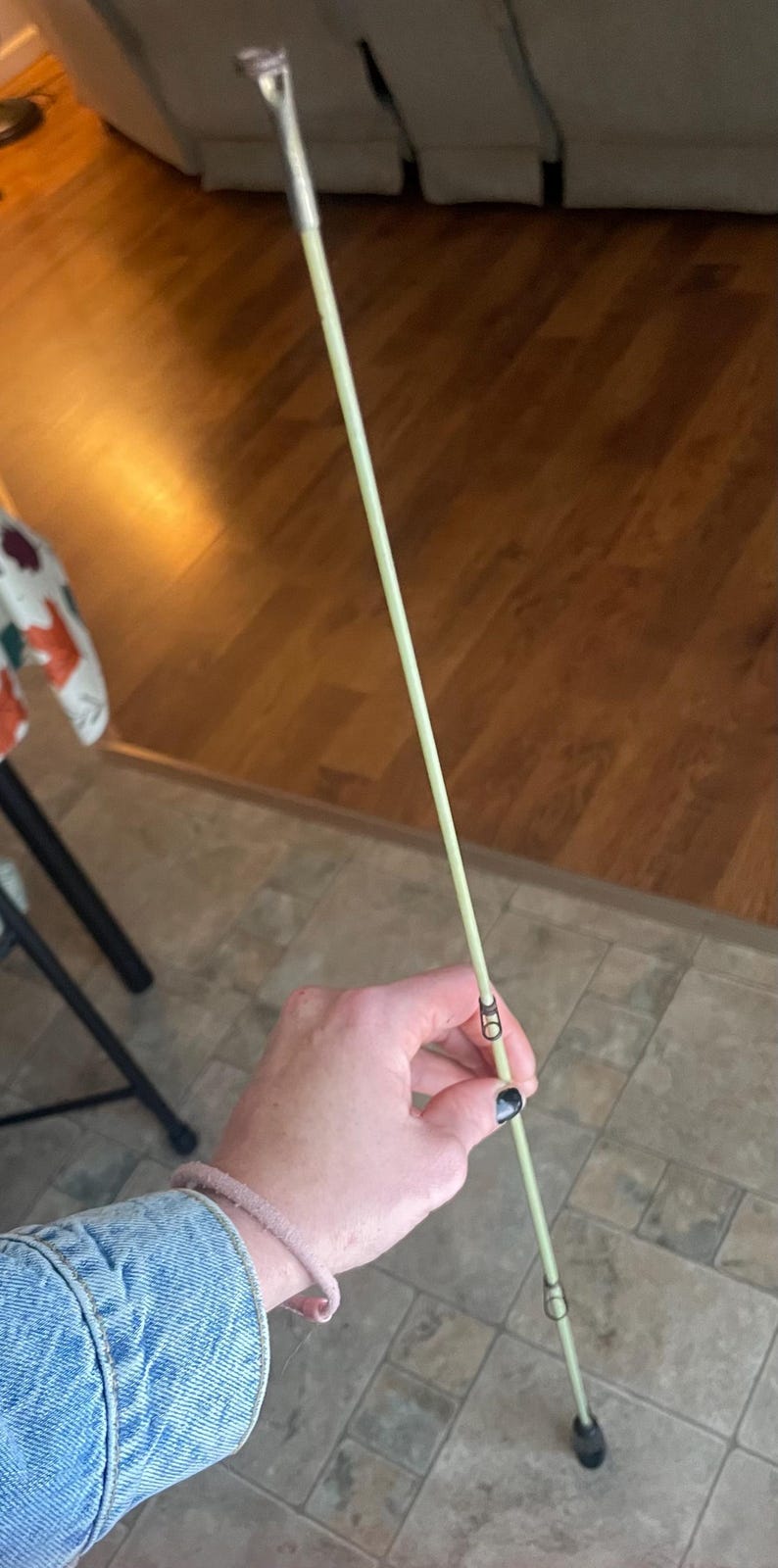 May include: A light green fishing rod with silver hardware. The rod is held at an angle, showing the guides and tip. The background is a wooden floor and a person's arm.