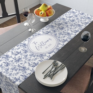May include: A blue and white table runner with a floral and rabbit design. The runner has an oval design with the text "The Williams EST. 2025". The table is set with plates, silverware, and wine glasses.