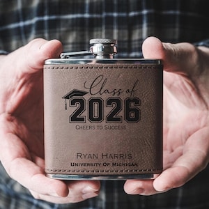 May include: A brown leather flask with a silver cap, engraved with "Class of 2026" and "Cheers to Success." The flask also features the name "Ryan Harris" and "University of Michigan."