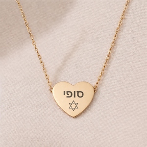 May include: A gold-colored necklace with a heart-shaped pendant. The pendant has the Hebrew word "סופי" and a Star of David symbol engraved in black. The necklace is displayed on a light-colored fabric.
