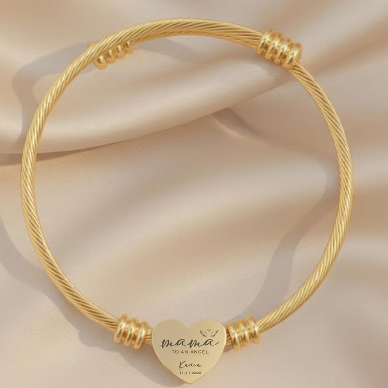 May include: Gold-colored bangle bracelet with a heart-shaped charm. The charm is engraved with the words "mama to an angel" and a name and date. The bracelet has a twisted cable design and is a piece of jewelry.