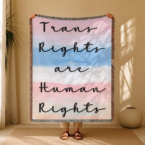 Könnte beinhalten: Eine gewebte Decke mit einem rosa, weißen und blauen Aquarelldesign. Die Worte "Trans Rights are Human Rights" sind in schwarzer Schreibschrift über die Decke geschrieben. Die Decke hat einen Fransenrand.