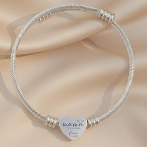 May include: A silver-colored wire bracelet with a heart-shaped charm. The charm has the words "mama to an angel" and a name and date. The bracelet is on a beige satin surface.