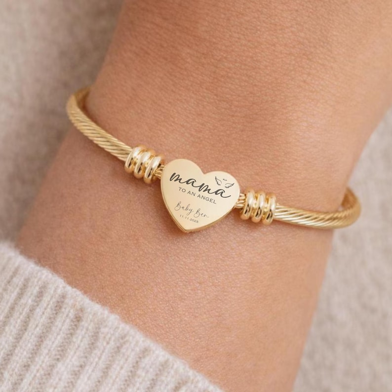 May include: Gold-toned bangle bracelet with a heart-shaped charm. The charm is engraved with the words "mama to an angel" and "Baby Bee" along with a date. The bracelet has a twisted cable design and is secured with decorative metal accents.