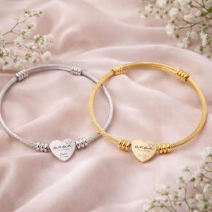 May include: Two bangle bracelets, one silver-toned and one gold-toned, each featuring a heart-shaped charm. The charms are engraved with the word "mama" and other text. The bracelets are displayed on a pink fabric background.