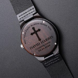 May include: A wooden wristwatch with a dark brown face and black leather strap. The watch face features a cross design and the engraved text: "MAY YOUR FAITH ALWAYS GUIDE YOU" and "DAVID HARRIS CONFIRMATION 03.26.2026".